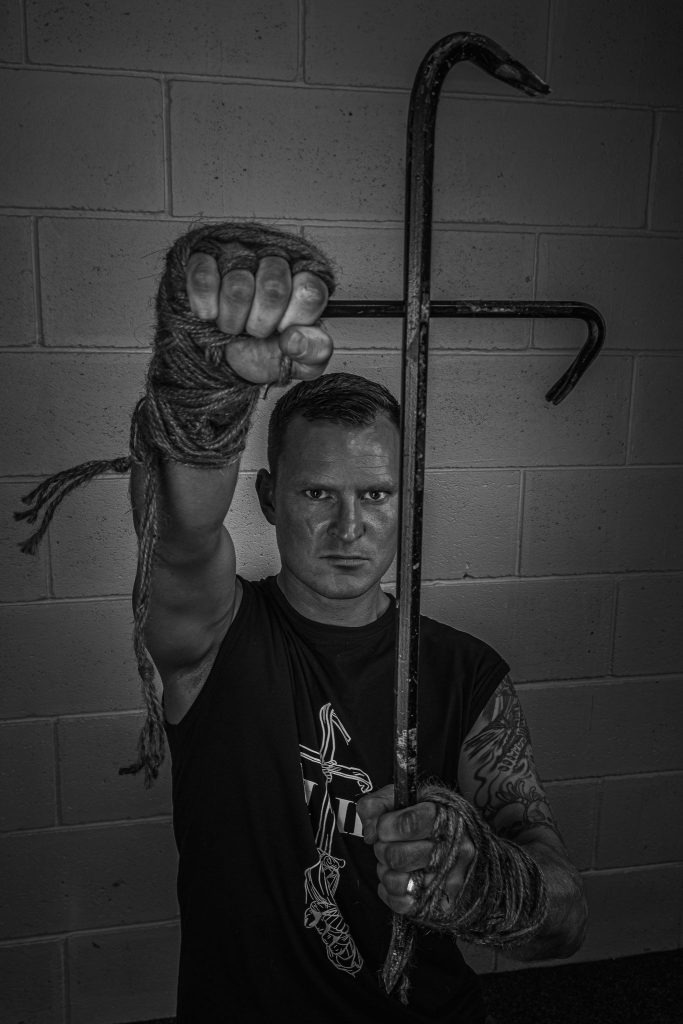 A black and white photo of a male wrestler kneeling on the floor, holding two crowbars to make a cross