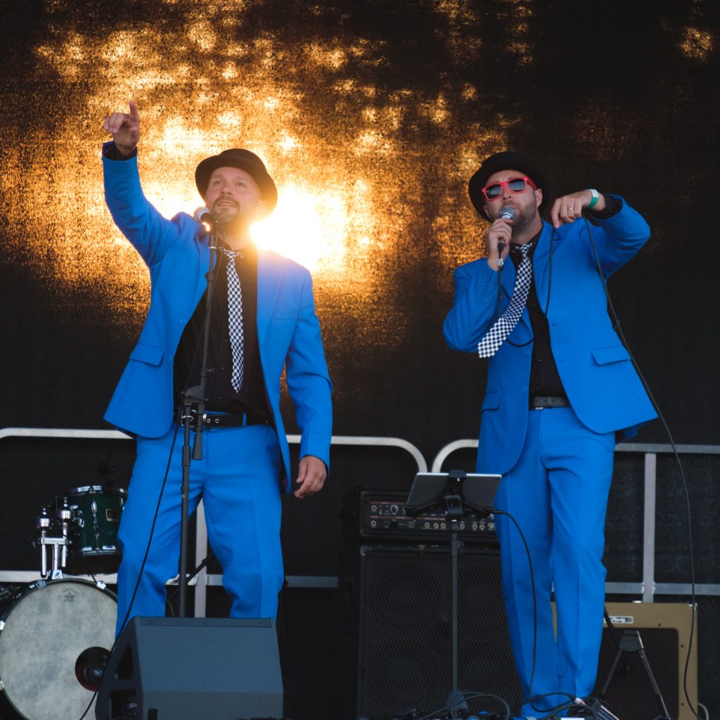 Two men dressed in blue suits on stage in front of a sun-lit backdrop