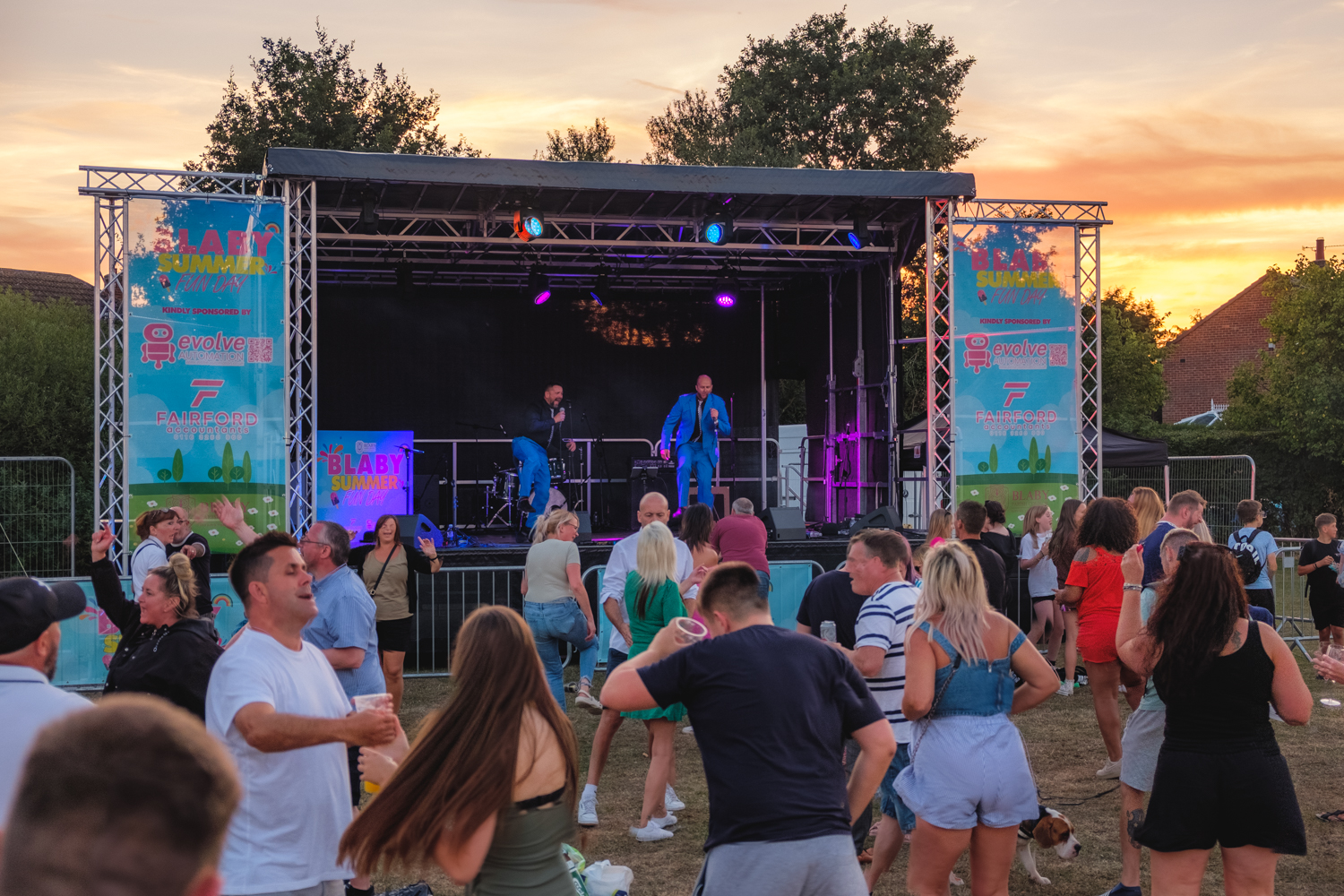 Two men in suits on a stage, with a sunset backdrop and a crowd of all types of people dancing